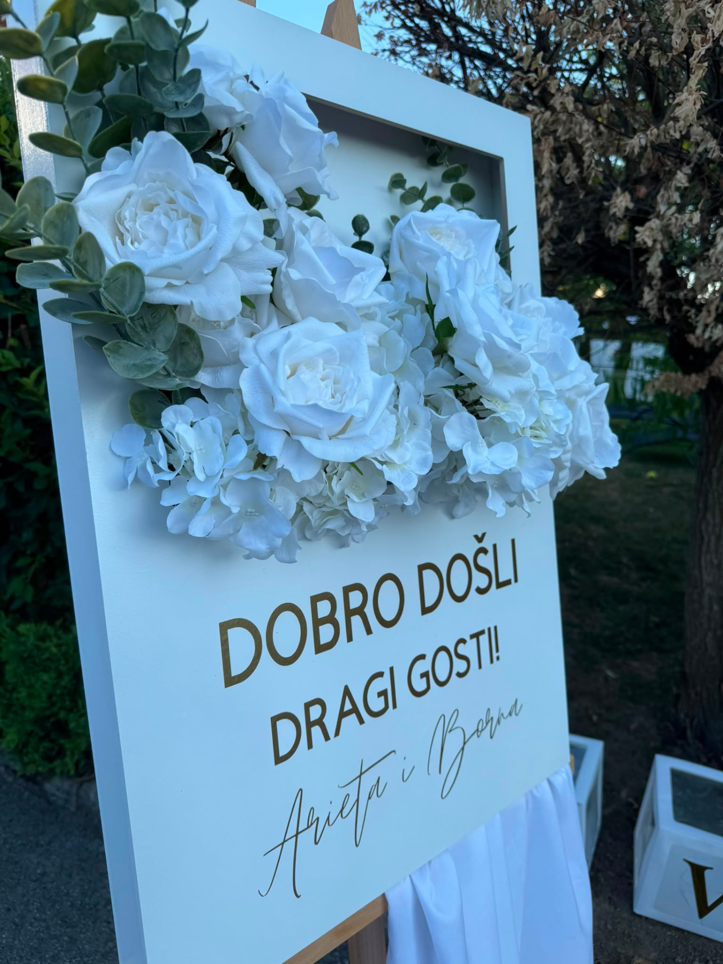 Bijela welcome ploča s bijelim ružama i hortenzijama te zlatnim natpisom Dobro došli dragi gosti